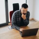 Young man with beard working on laptop at desk in a modern office setting.